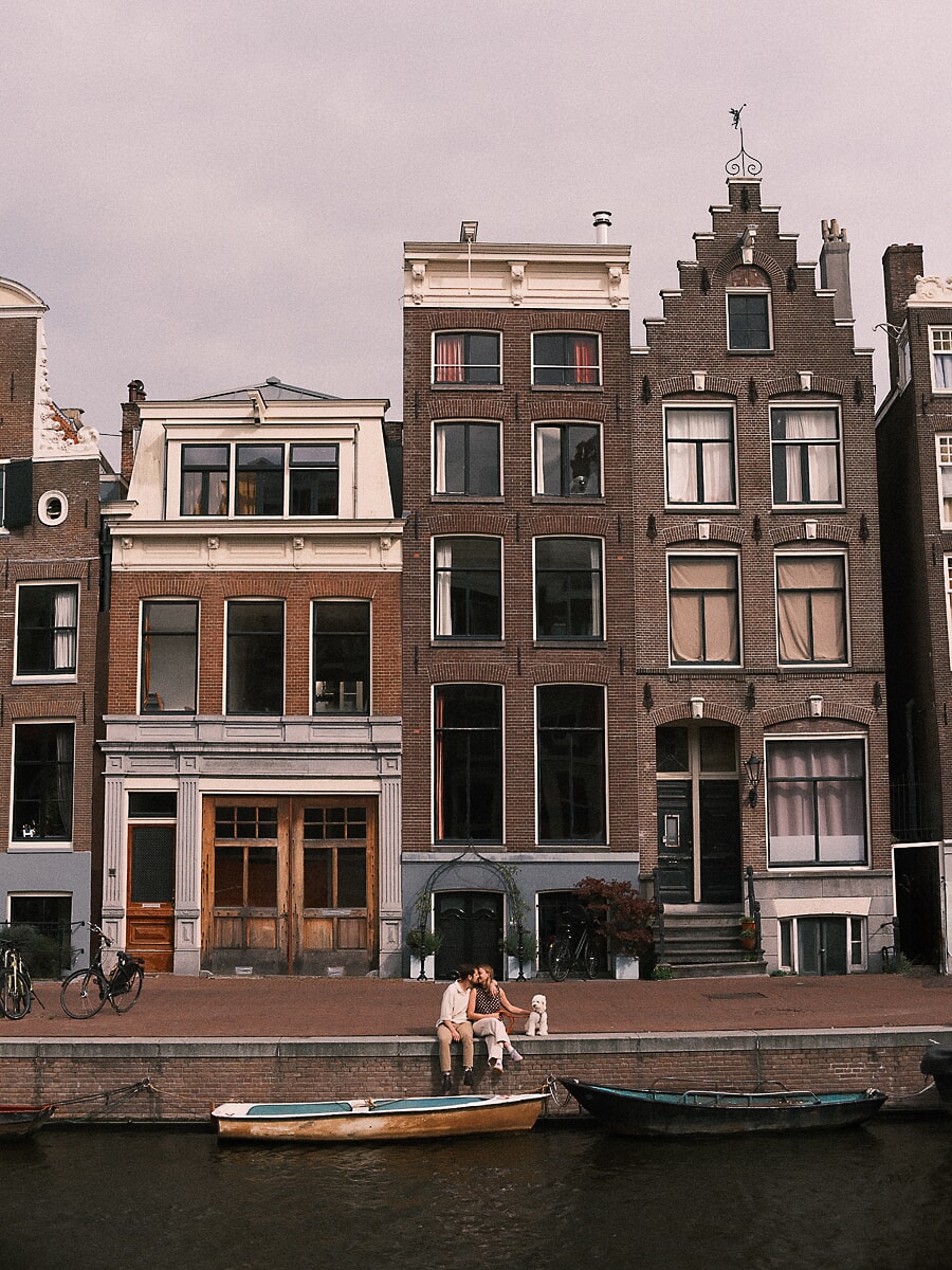 Engagement SP5 Breed gekaderde koppelfotografie in Amsterdam met een stel dat elkaar kust aan de gracht voor de trapgevels