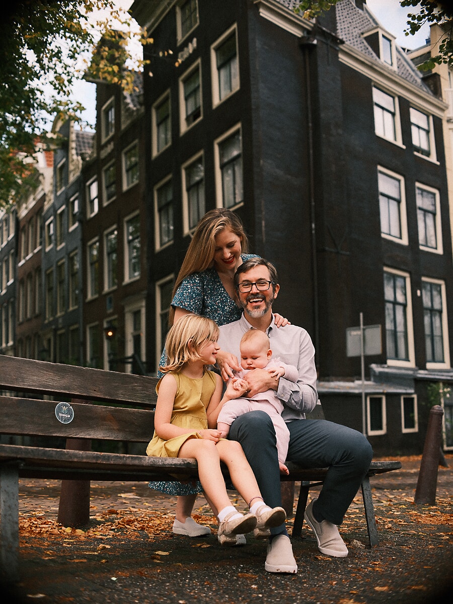 Family SP10 Familie van vier op een houten bank tussen de gevallen bladeren bij een gracht, het oudste meisje in het geel naast haar baby, vastgelegd met cinematische familiefotografie
