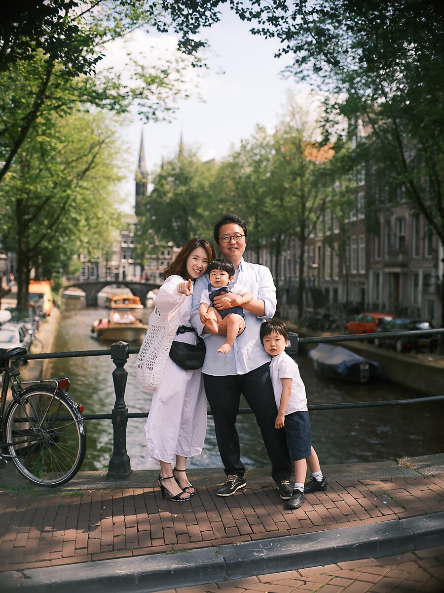 Family SP12 Expatgezin op een brug over een gracht in de zomer tijdens een familiefotoshoot in Amsterdam, de ouders met een peuter naast hun jonge zoon