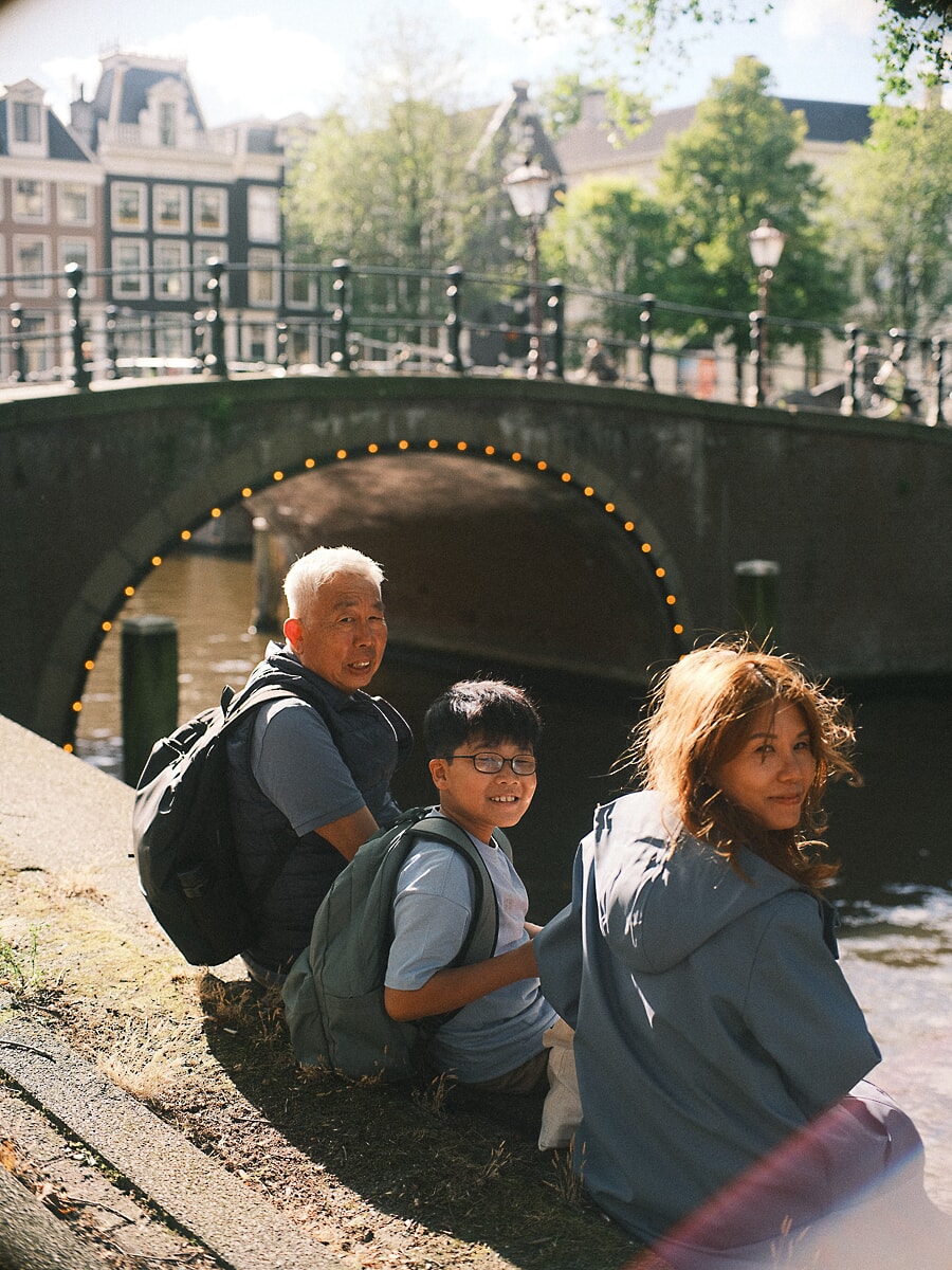 Family SP13 Drie generaties gehurkt bij een gracht onder een verlichte boogbrug, vastgelegd door een familiefotograaf in Amsterdam
