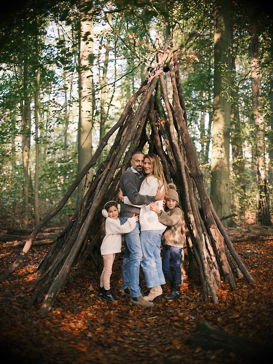 Family SP14 Een gezin van vier omhelst elkaar in een tipi van takken in het herfstbos, een sfeervol moment vol familiefotografie