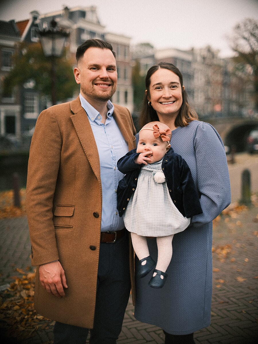 Family SP16 Kersverse ouders met hun baby op een brug over de gracht in de herfst, een portret door een familiefotograaf in Amsterdam, de vader in een kameelkleurige jas