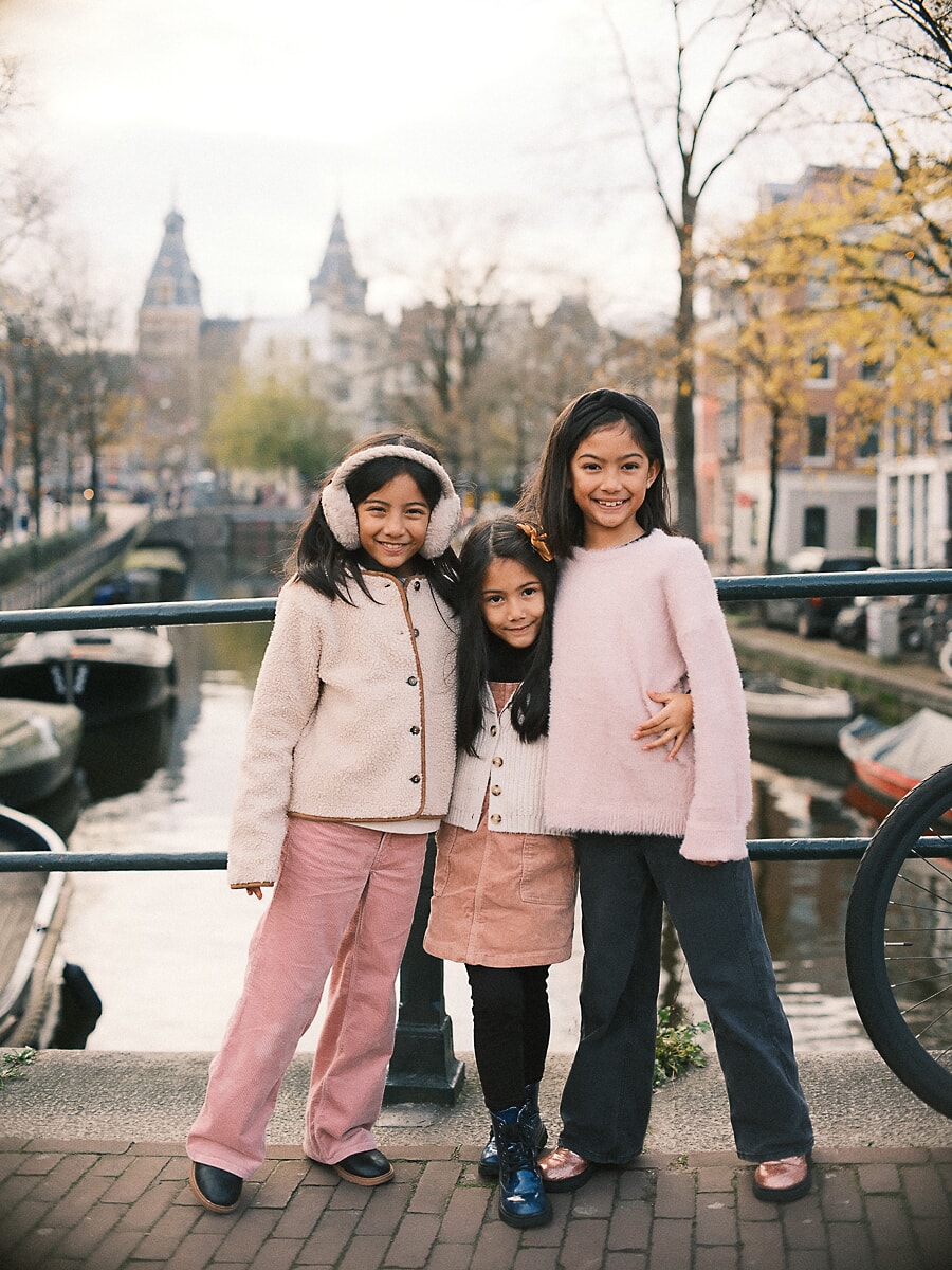 Family SP4 Zusjes in roze en crème poseren bij een reling langs een Amsterdamse gracht, een opgewekt beeld uit een kinderfotoshoot