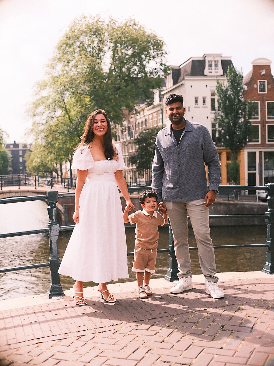 Family SP7 Lachende ouders houden de handen van hun jonge zoon vast op een zonovergoten Amsterdamse brug, de moeder in een witte jurk tijdens een gezinsshoot in de zomer