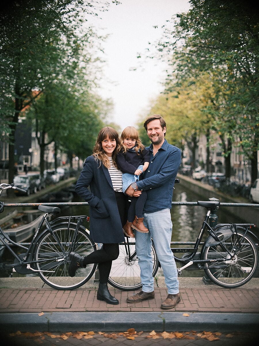 Family SP8 Familiefotograaf in Amsterdam legt ouders met hun peuter vast naast fietsen langs de gracht op een herfstmiddag