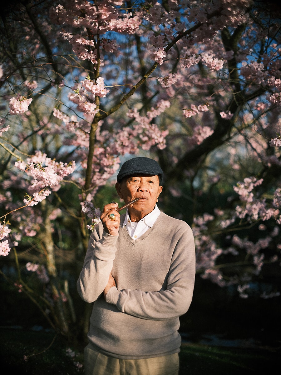 Group Family SP2 Expressief portret van een opa met pet die even stilstaat onder bloeiende kersentakken, onderdeel van een familieshoot met meerdere generaties