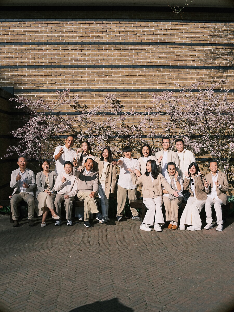 Group Family SP8 Grote familiefotoshoot met meerdere generaties in crèmetinten, familieleden van alle leeftijden voor een bakstenen muur en bloeiende kersenboom