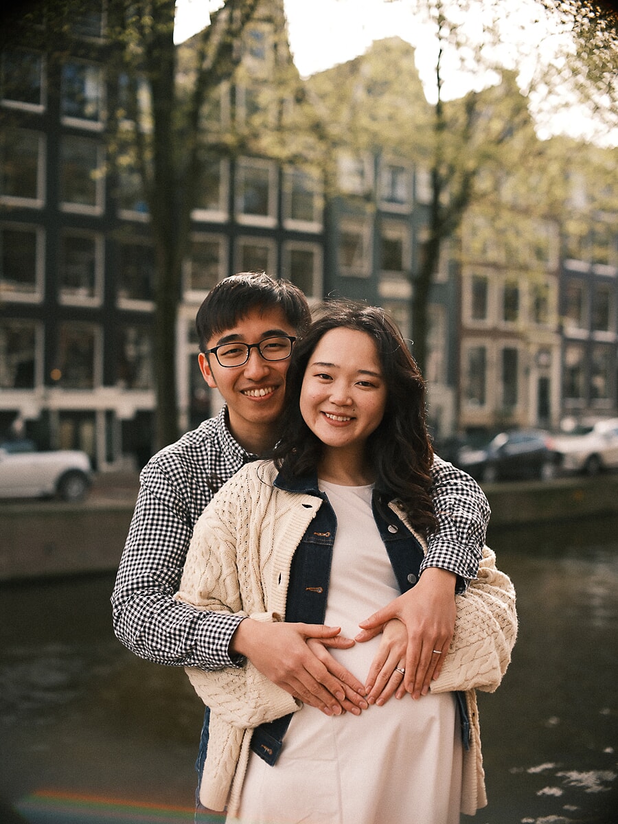 Maternity SP1 Lachend koppel bij een Amsterdamse gracht terwijl de partner zijn armen om de aanstaande moeder slaat en een hartje vormt op haar zwangere buik tijdens hun zwangerschapssessie