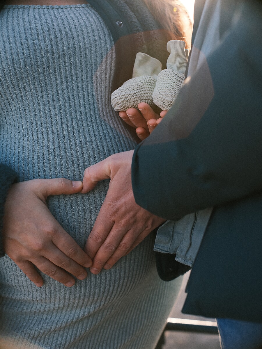 Maternity SP3 Tegenlichtopname tijdens een babybuikshoot: de handen van een koppel houden kleine gebreide babyschoentjes vast tegen een zwangere buik, de vrouw in een blauwe jurk