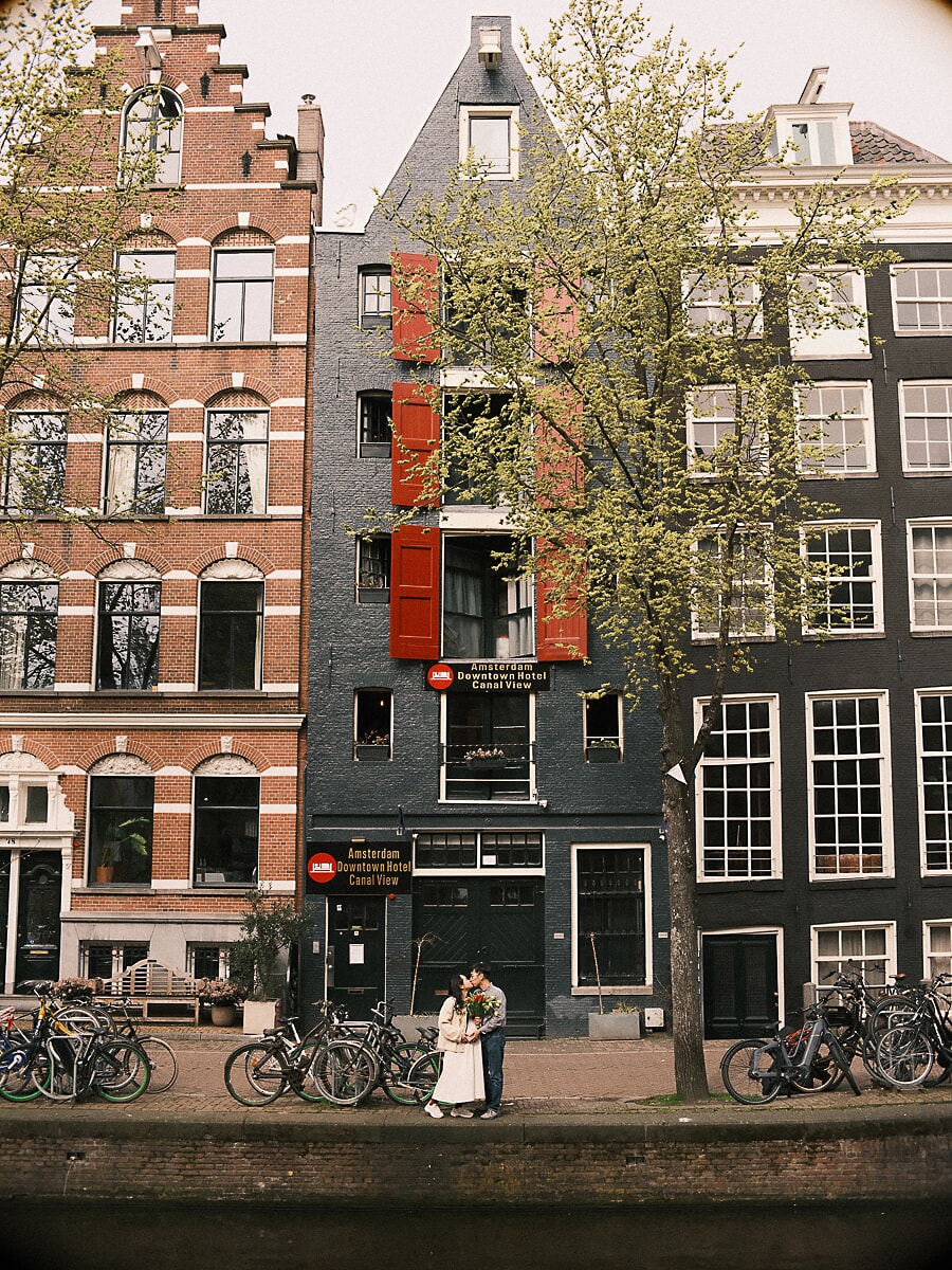 Maternity SP4 Hoge grachtenpanden met rode luiken torenen boven een zoenend stel tussen geparkeerde fietsen tijdens hun zwangerschapsfotoshoot in Amsterdam
