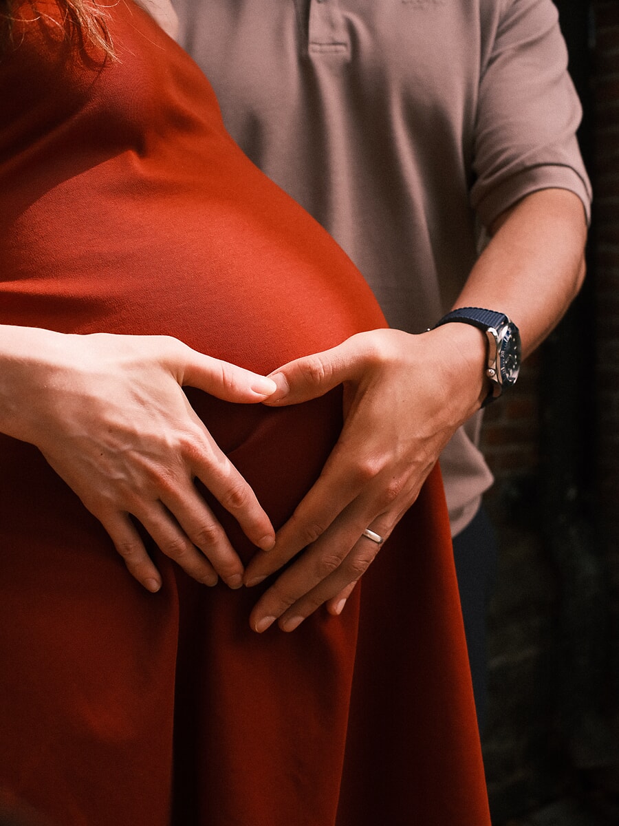 Maternity SP5 Intieme zwangerschapsfoto's van een koppel dat met hun handen een hartje vormt op de babybuik, de zwangere vrouw in een roestkleurige jurk