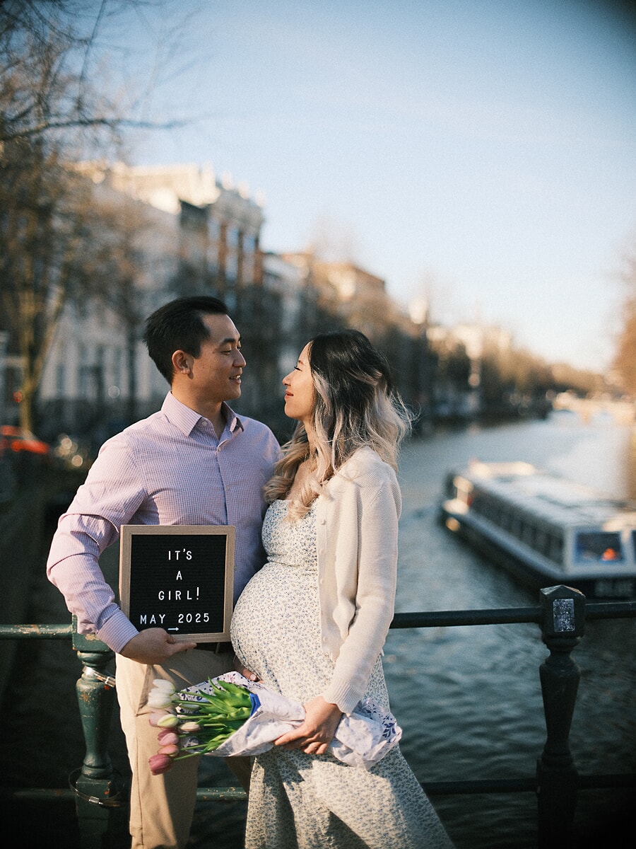 Maternity SP6 Genderreveal-fotoshoot bij een Amsterdamse gracht, waar een koppel een 'It's a girl'-bord en een tulpenboeket vasthoudt, met een rondvaartboot op de achtergrond