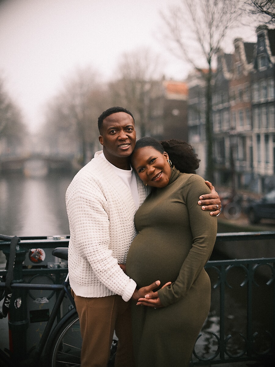 Maternity SP7 Vrolijke zwangerschapsportretten van een koppel dat samen de buik vasthoudt bij een reling langs de gracht, de aanstaande moeder in een olijfgroene jurk en haar partner in een crèmekleurige trui