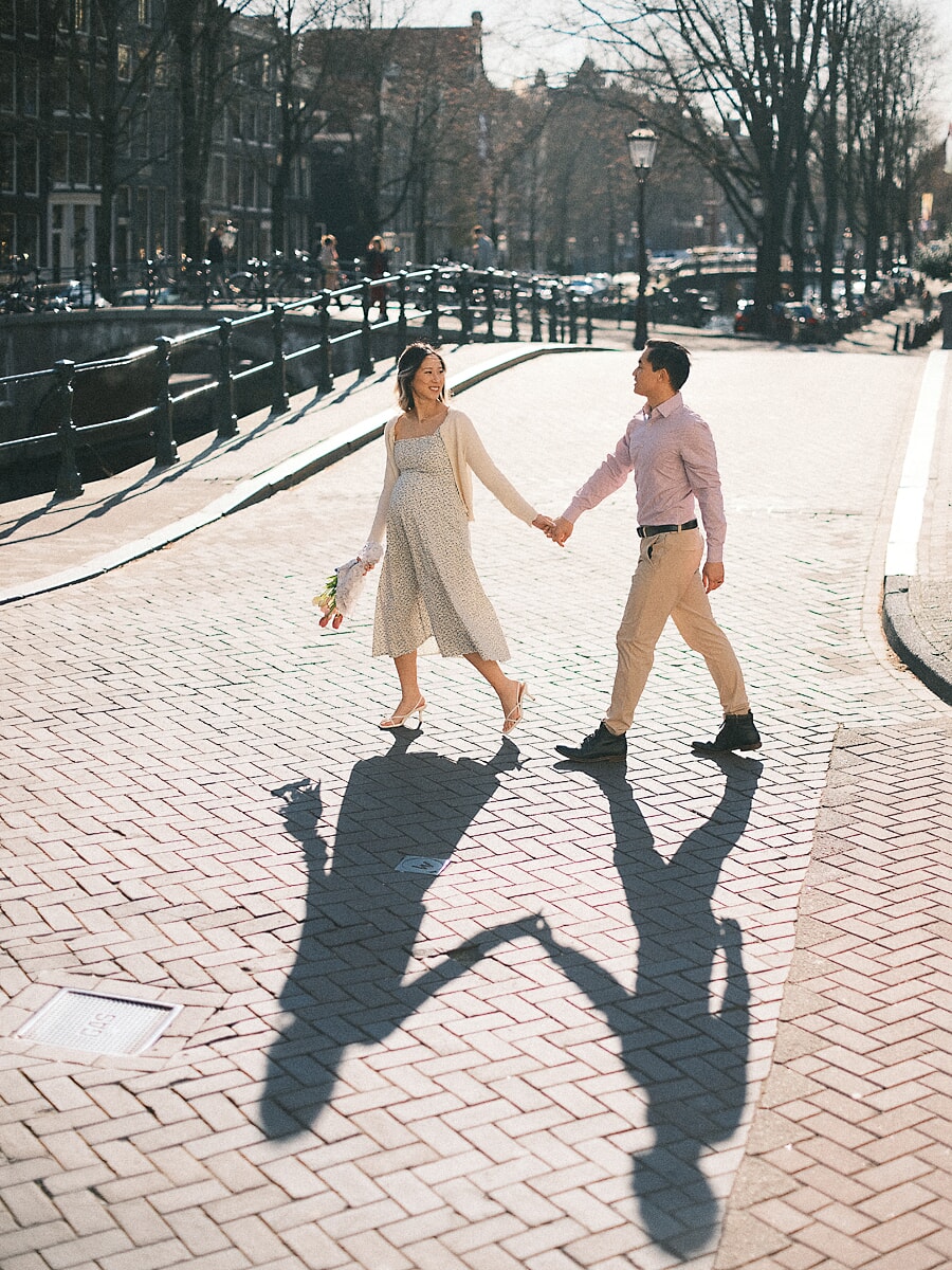 Maternity SP8 Zwangere vrouw en haar partner lopen hand in hand over een zonovergoten Amsterdamse brug tijdens hun babymoon, met lange schaduwen over de kasseien