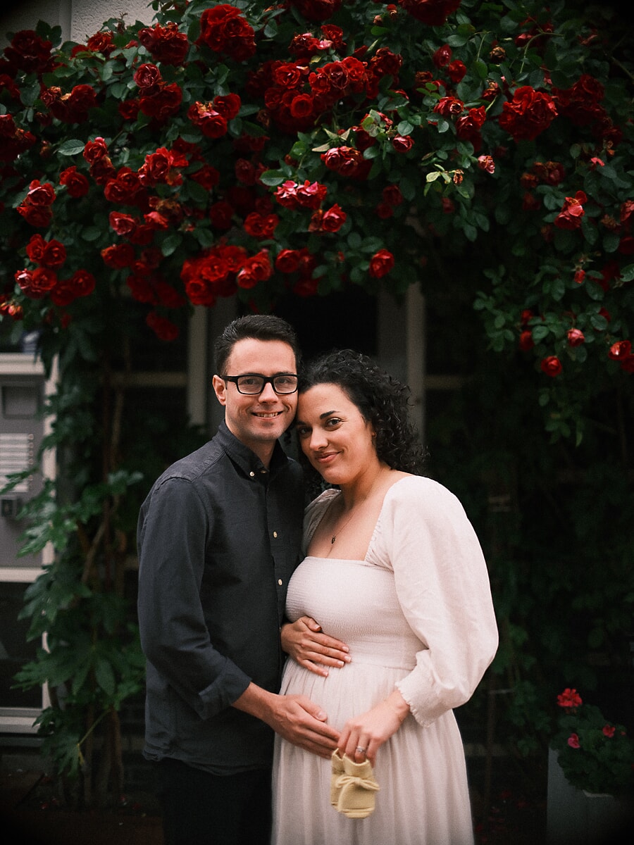 Maternity SP9 Aanstaande ouders tijdens een zwangerschapsshoot onder een muur van rode klimrozen, met kleine gebreide babyslofjes tegen de buik van de moeder