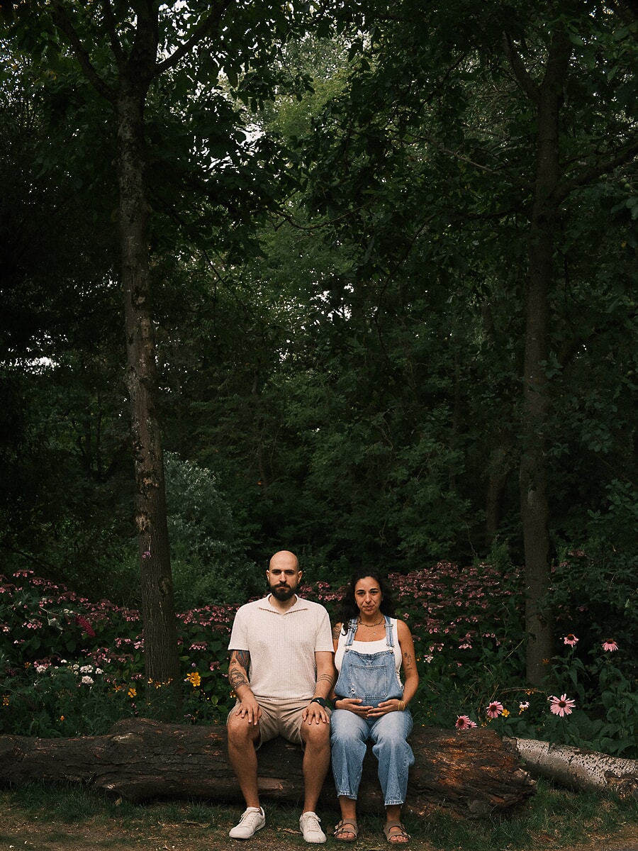 Maternity SP10 Ontspannen zwangerschapsfotoshoot van een koppel op een omgevallen boomstam in een weelderige bloementuin, de aanstaande moeder in een denim tuinbroek met een hand op haar babybuik