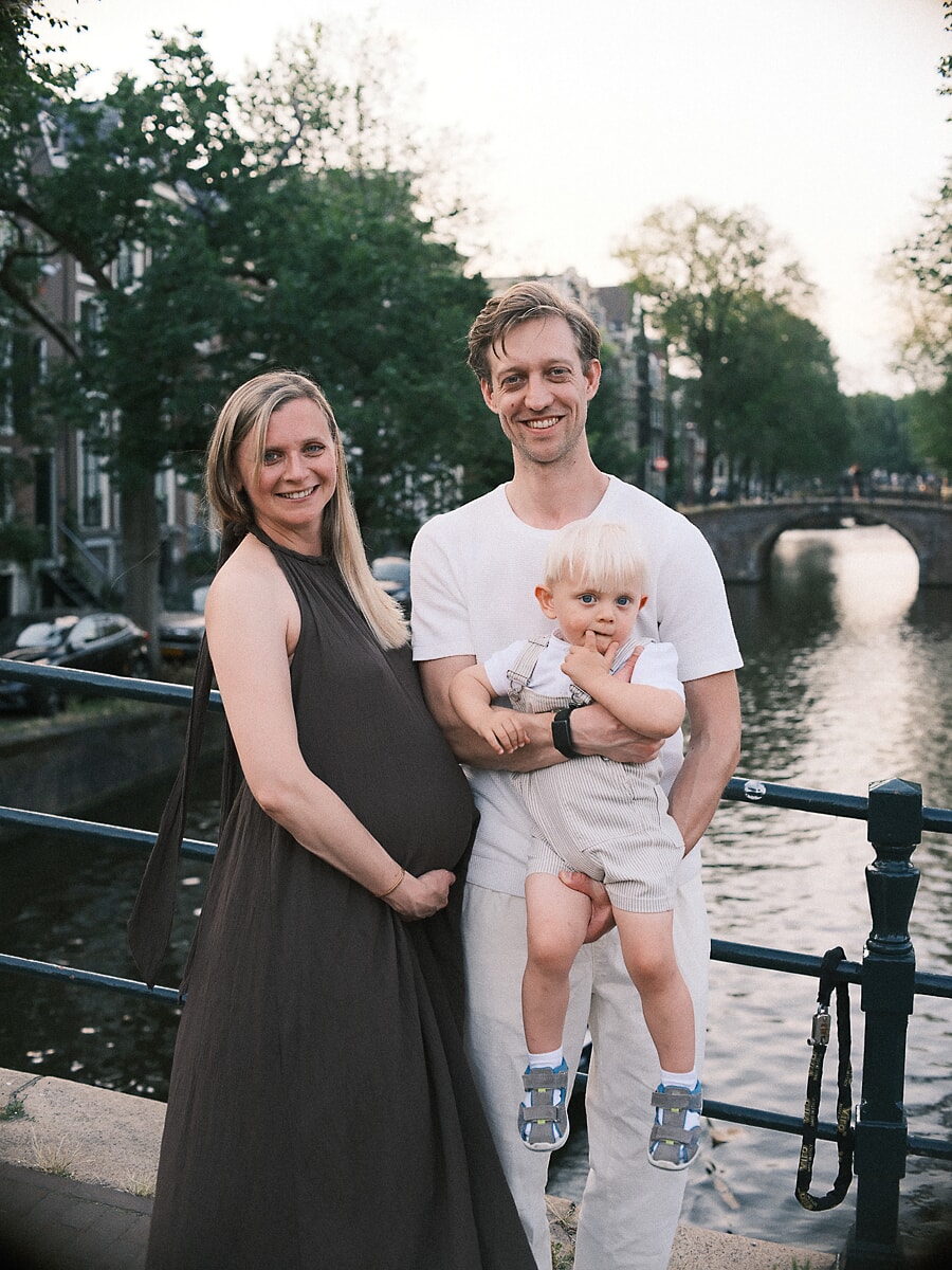 Maternity SP11 Groeiend gezin op een zonovergoten brug over de gracht tijdens een zomerse zwangerschapssessie, de vader met hun jonge zoon naast de aanstaande moeder in een donkere mouwloze jurk, met een boogbrug op de achtergrond