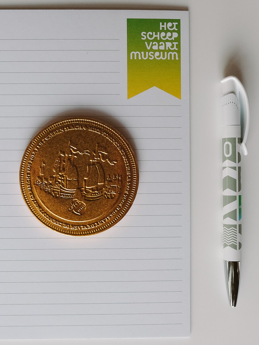 Flat-lay van een gelinieerd notitieblok, een gouden penning in reliëf en een bedrukte pen, productfotografie in Amsterdam