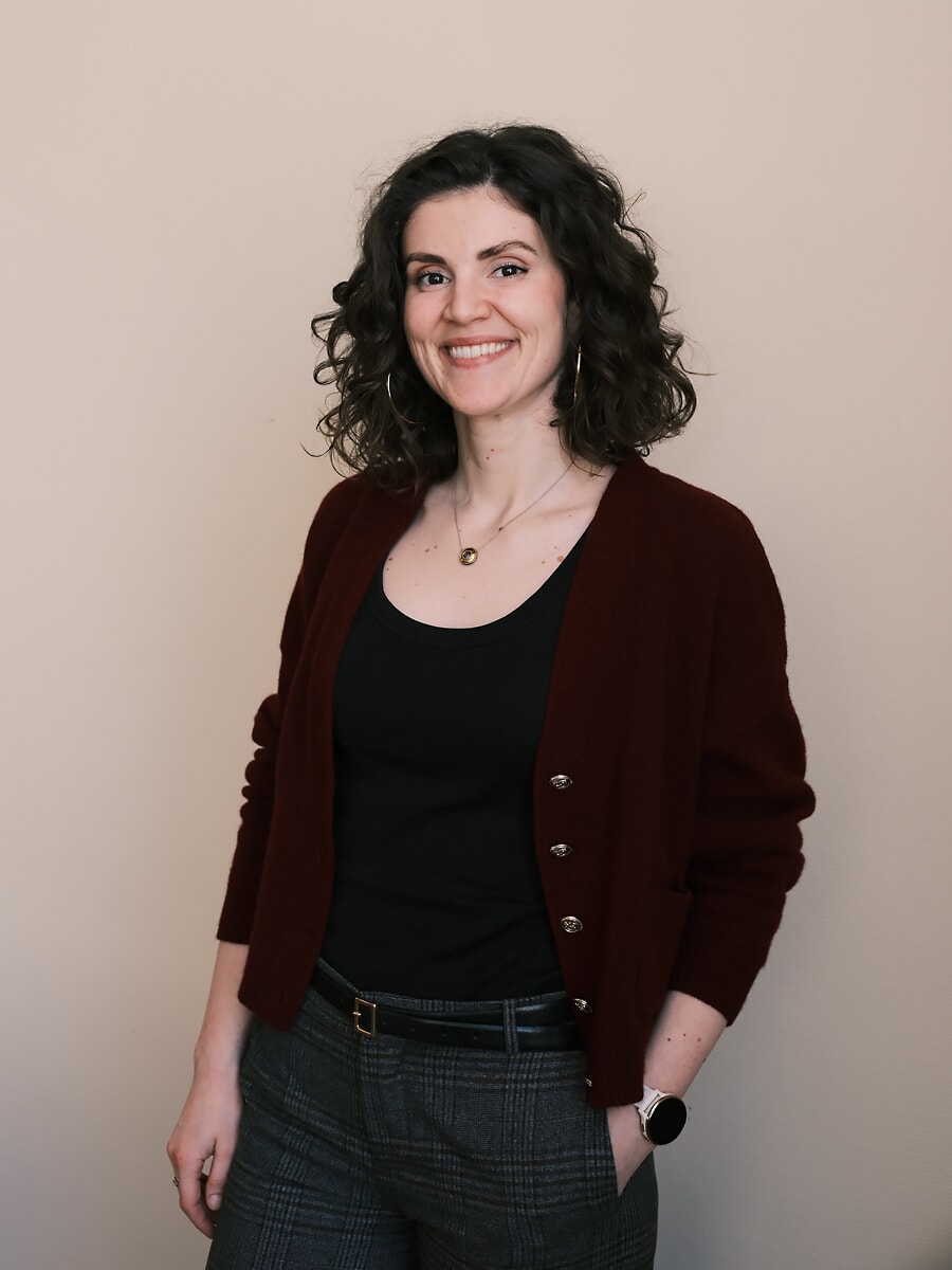 Vrouw met krullend haar in een bordeauxrood vest en geruite broek, een hand in haar zak tegen een beige muur