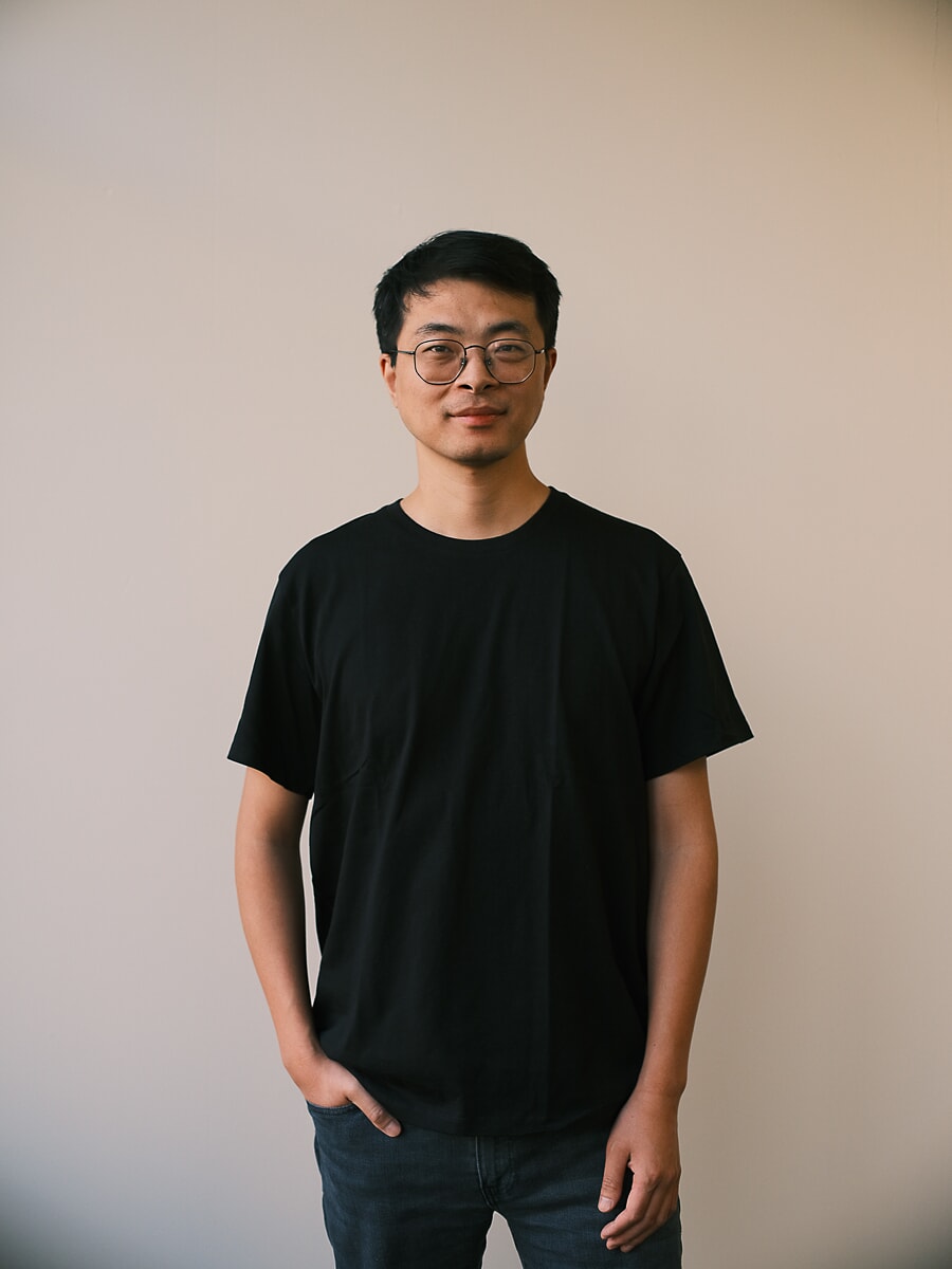 Man met ronde bril en zwart T-shirt, handen in zijn zakken tegen een effen lichte muur, een zakelijk portret in Amsterdam