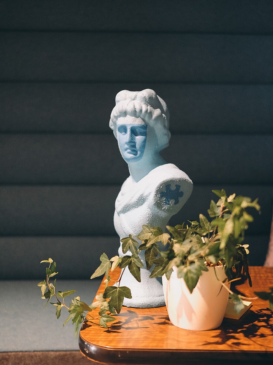 Lichtblauwe klassieke buste en een pot klimop op een houten tafel tegen een donkere geribbelde bank, met zonlicht eroverheen