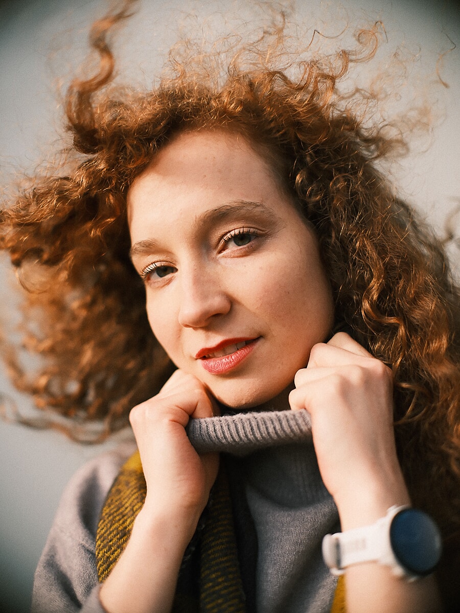 Close-up van een vrouw met rode krullen die een grijze coltrui tot haar kin optrekt tegen een open lucht