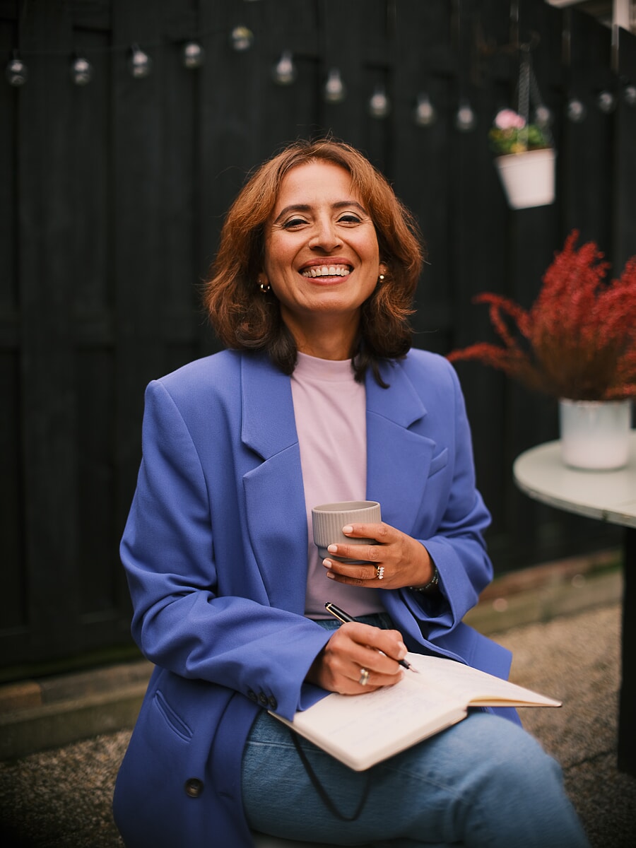 Coach in een blauwe blazer lacht met een kop koffie en een open notitieboek onder lichtjes, een brandingshoot in Amsterdam