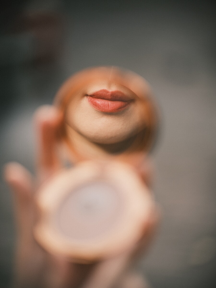 Extreme close-up van een hand met een rond spiegeltje waarin de lippen van een vrouw weerspiegelen, de rest zacht en onscherp