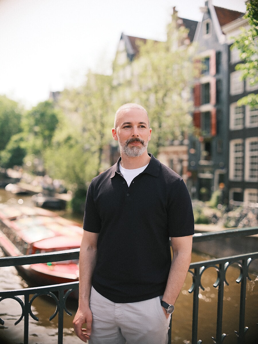 Man met grijze baard in donkerblauwe polo bij een reling langs de gracht met afgemeerde boten en trapgevels erachter, een confidenceportret in Amsterdam