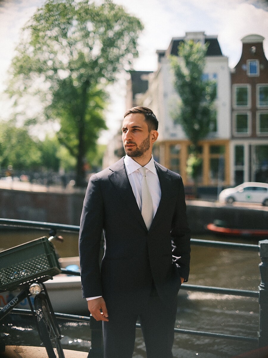 Man in donkerblauw pak met stropdas kijkt langs een reling aan de gracht, met lichte trapgevels achter hem, een formeel confidenceportret in Amsterdam