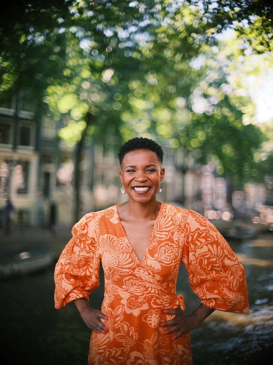 Vrouw in oranje jurk met bladprint lacht met haar handen in haar zij, met gefilterd grachtlicht achter zich, een levendig confidenceportret