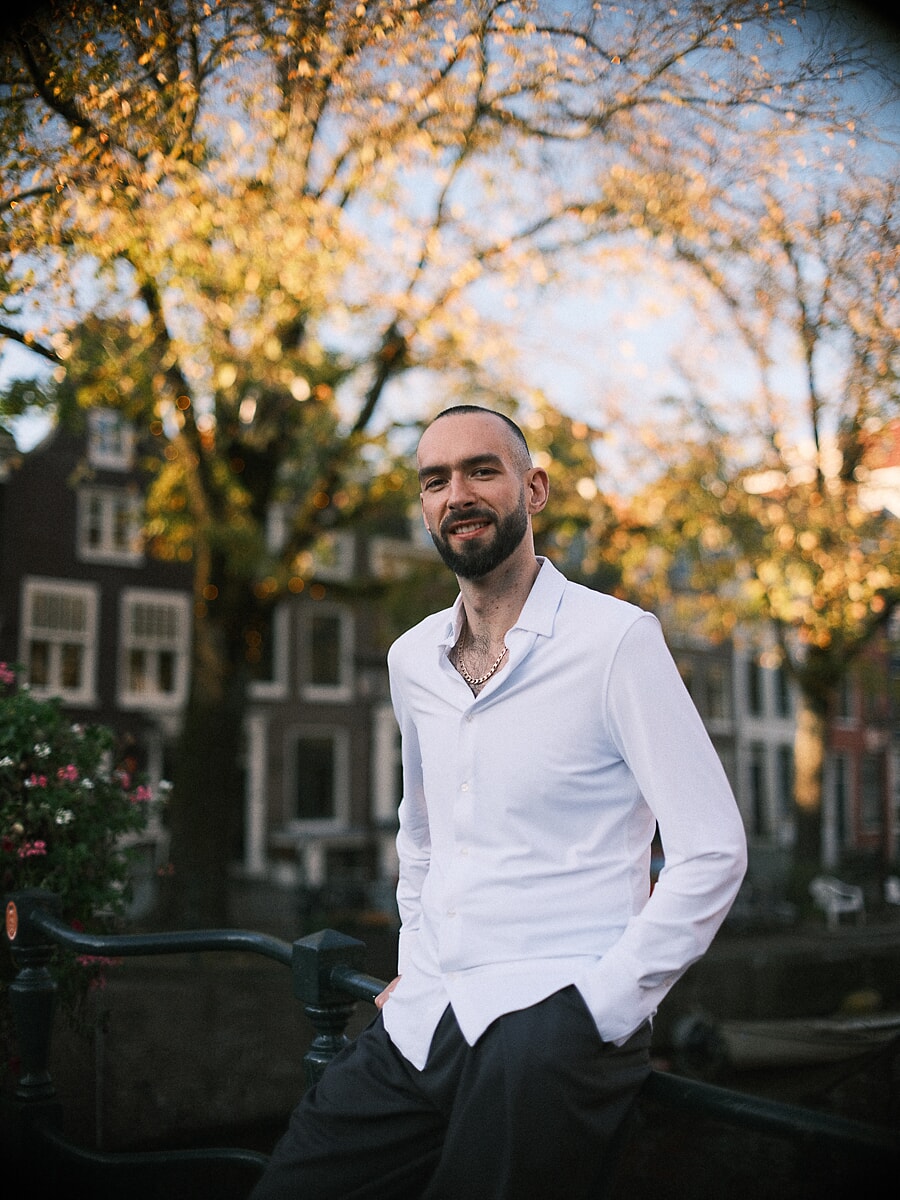 Man in wit overhemd leunt tegen een reling langs de gracht onder gouden herfstbladeren, een warm confidenceportret in Amsterdam