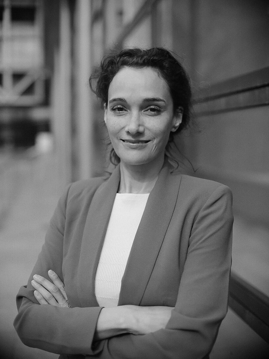 Vrouw met opgestoken haar en gekruiste armen in een lichte blazer, een industriële constructie onscherp erachter, een zakelijk portret in zwart-wit