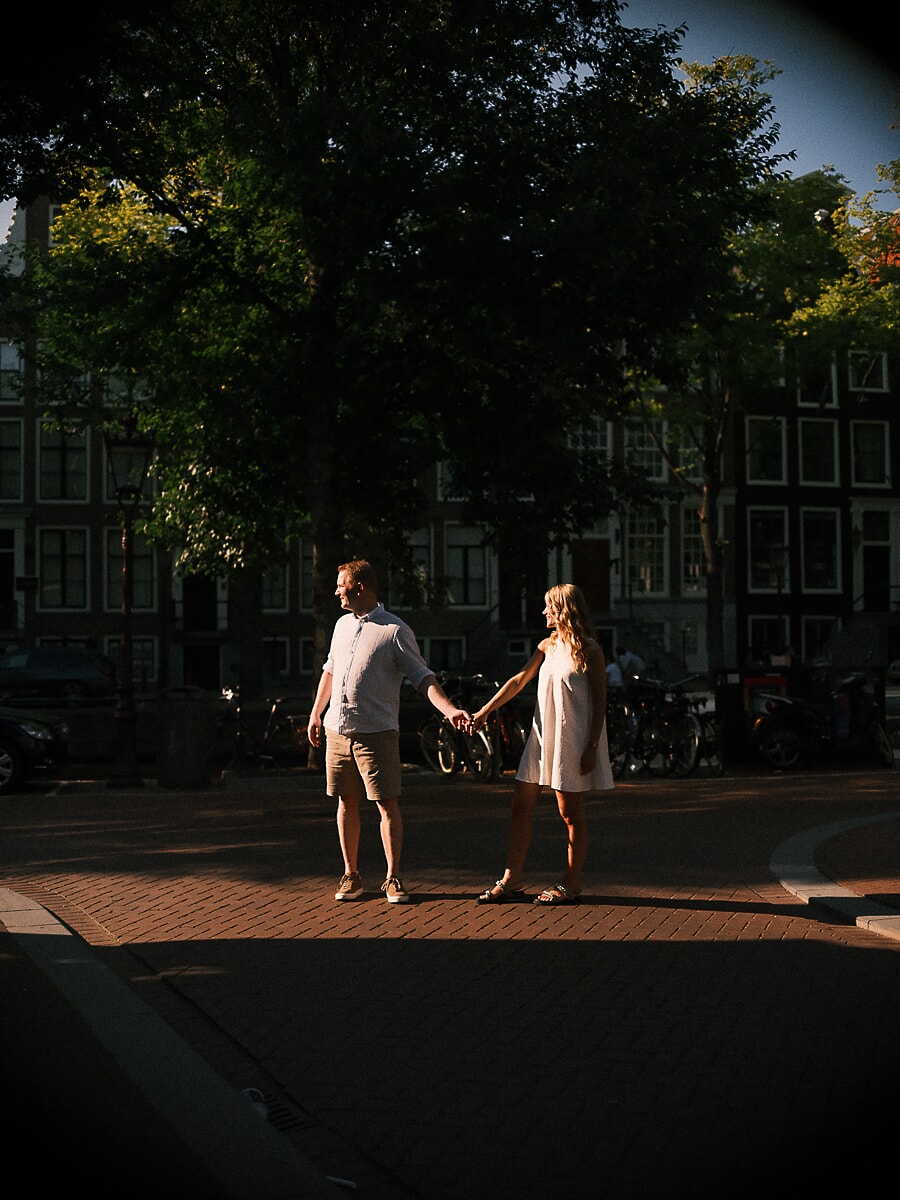 Stel houdt elkaars handen op armlengte vast in een strook avondlicht, een spontaan beeld op straat