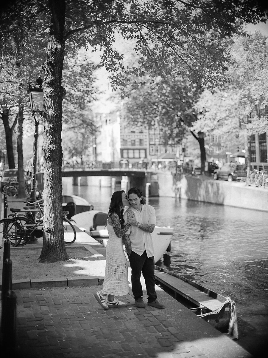 Zwart-witbeeld van een stel op een geplaveide kade in Amsterdam, zij kust zijn wang naast de afgemeerde boten