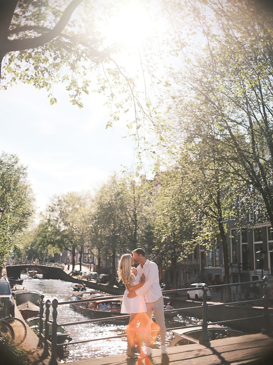 Stel in het wit kust bij een grachtenhek onder hoge zonovergoten bomen, met een warme lensgloed tijdens hun verlovingsfotoshoot in Amsterdam
