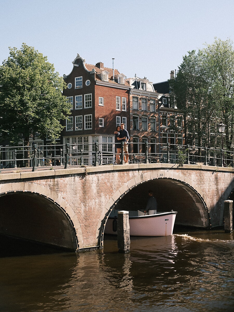 Stel omhelst elkaar op een stenen grachtbrug terwijl een boot onder hen doorvaart tijdens hun loveshoot in Amsterdam