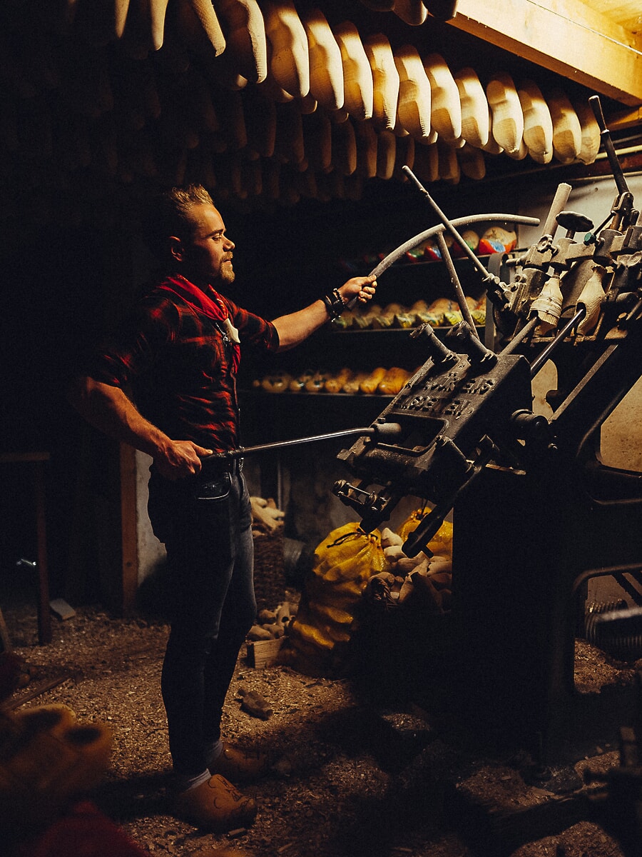 Vakman bedient een gietijzeren klompenmachine, met rijen houten klompen aan de balken boven hem