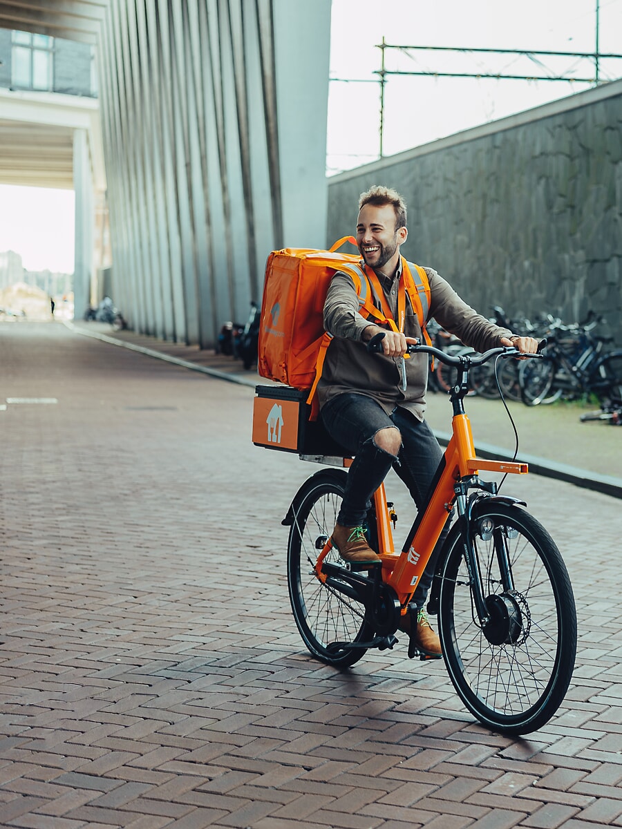 Bezorger lacht op een oranje e-bike met een geïsoleerde rugtas, op een klinkerstraat onder een modern gebouw in Amsterdam