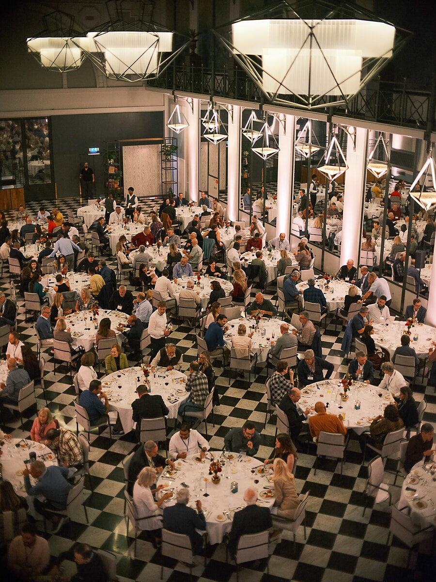 Eetzaal vol gasten, van bovenaf gezien, ronde tafels op een schaakbordvloer onder geometrische lampen, eventfotografie in Amsterdam