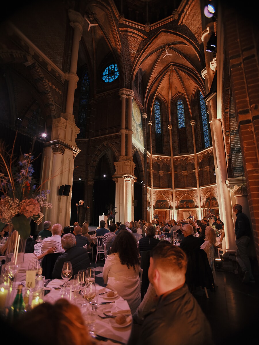 Diner in het schip van een gotische kerk, een spreker in de verte onder bakstenen gewelven en glas-in-lood, een bedrijfsevenement in Amsterdam