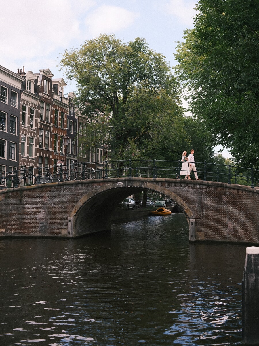 Verloofd stel steekt een gebogen Amsterdamse grachtbrug over, breed in beeld vanaf het water