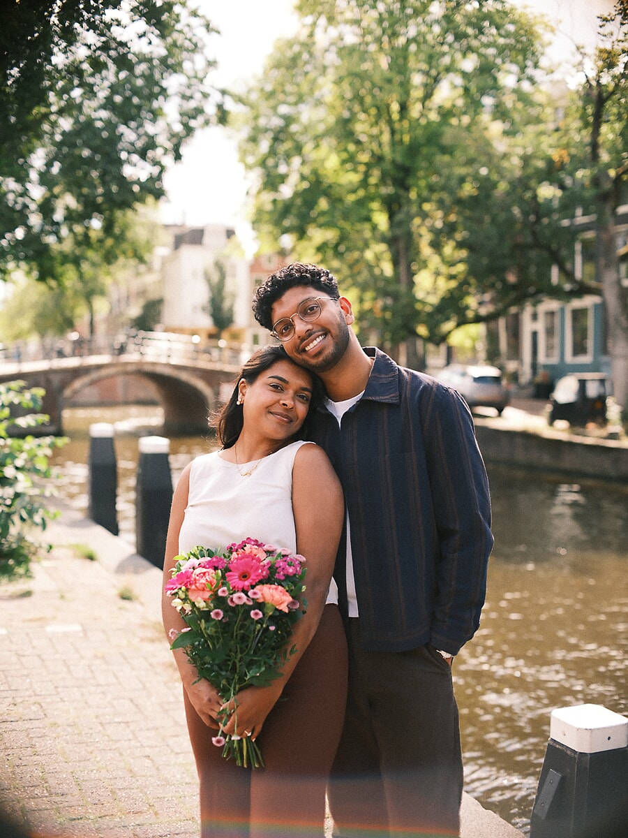 Verloofd stel dicht bij elkaar langs een Amsterdamse gracht met roze gerbera's en een gebogen brug erachter