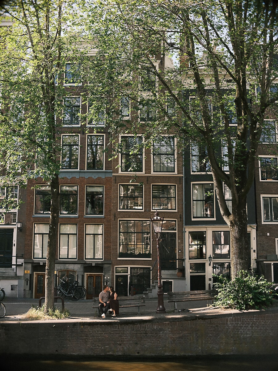 Verloofd stel klein tussen de hoge Amsterdamse grachtenpanden, een breed beeld uit hun verlovingsshoot