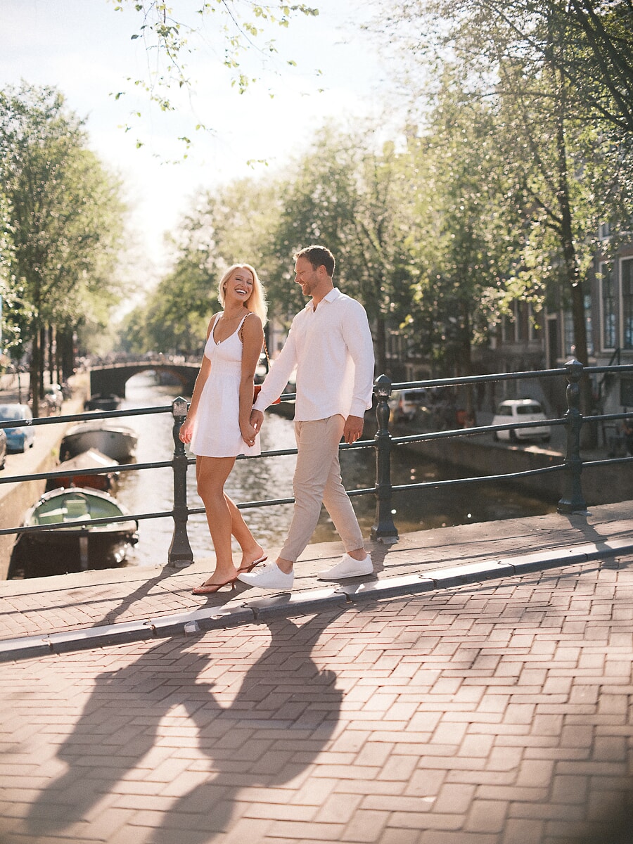 Pas verloofd stel loopt hand in hand over een zonnige kade in Amsterdam, zij kijkt lachend over haar schouder