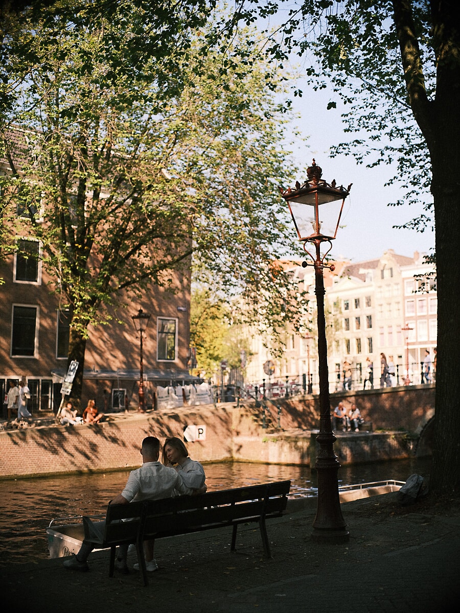 Verloofd stel zit dicht tegen elkaar op een bankje langs de gracht onder een Amsterdamse straatlantaarn