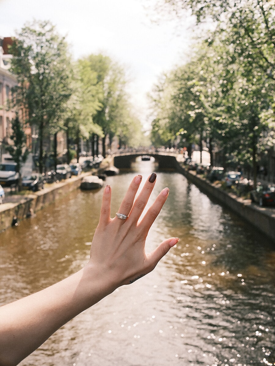 Verlovingsring aan een opgeheven hand boven een zonovergoten Amsterdamse gracht na het huwelijksaanzoek