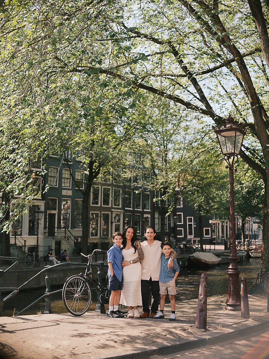 Ouders en hun twee zoons arm in arm op een zonnige kade in Amsterdam, met een sierlijke lantaarnpaal en hoge grachtenpanden