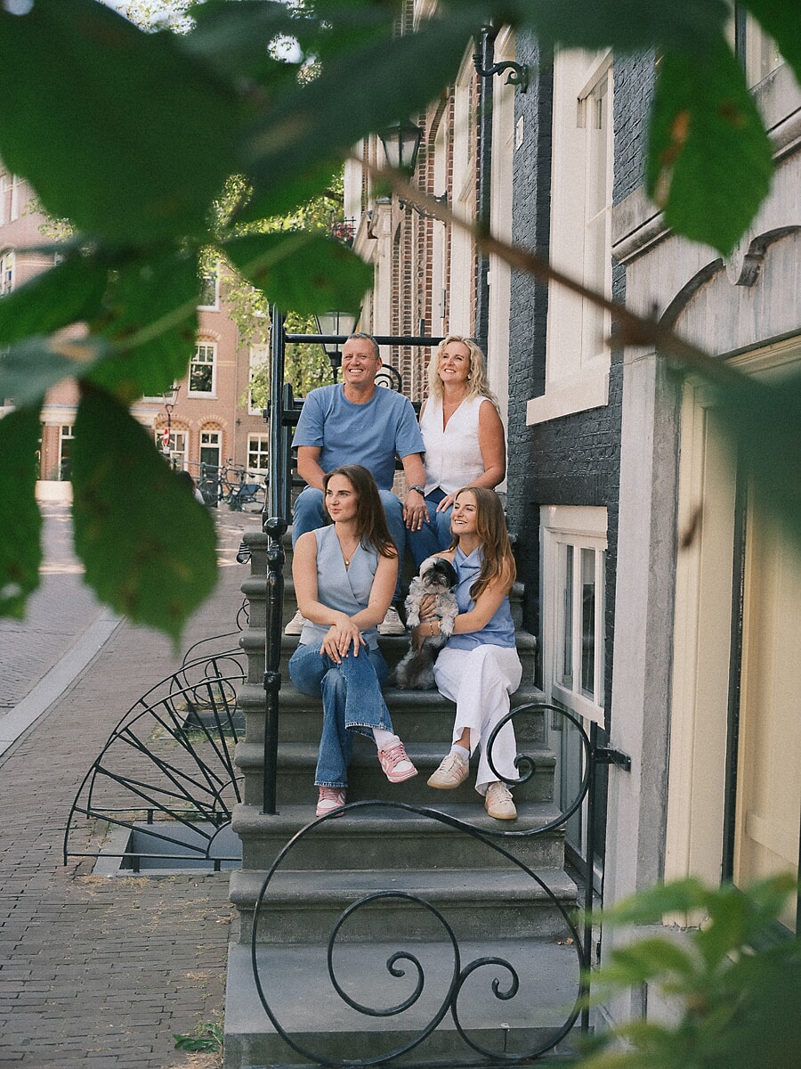 Gezin van vier met hun hondje op de stoep van een Amsterdams grachtenpand, gezien door groene bladeren