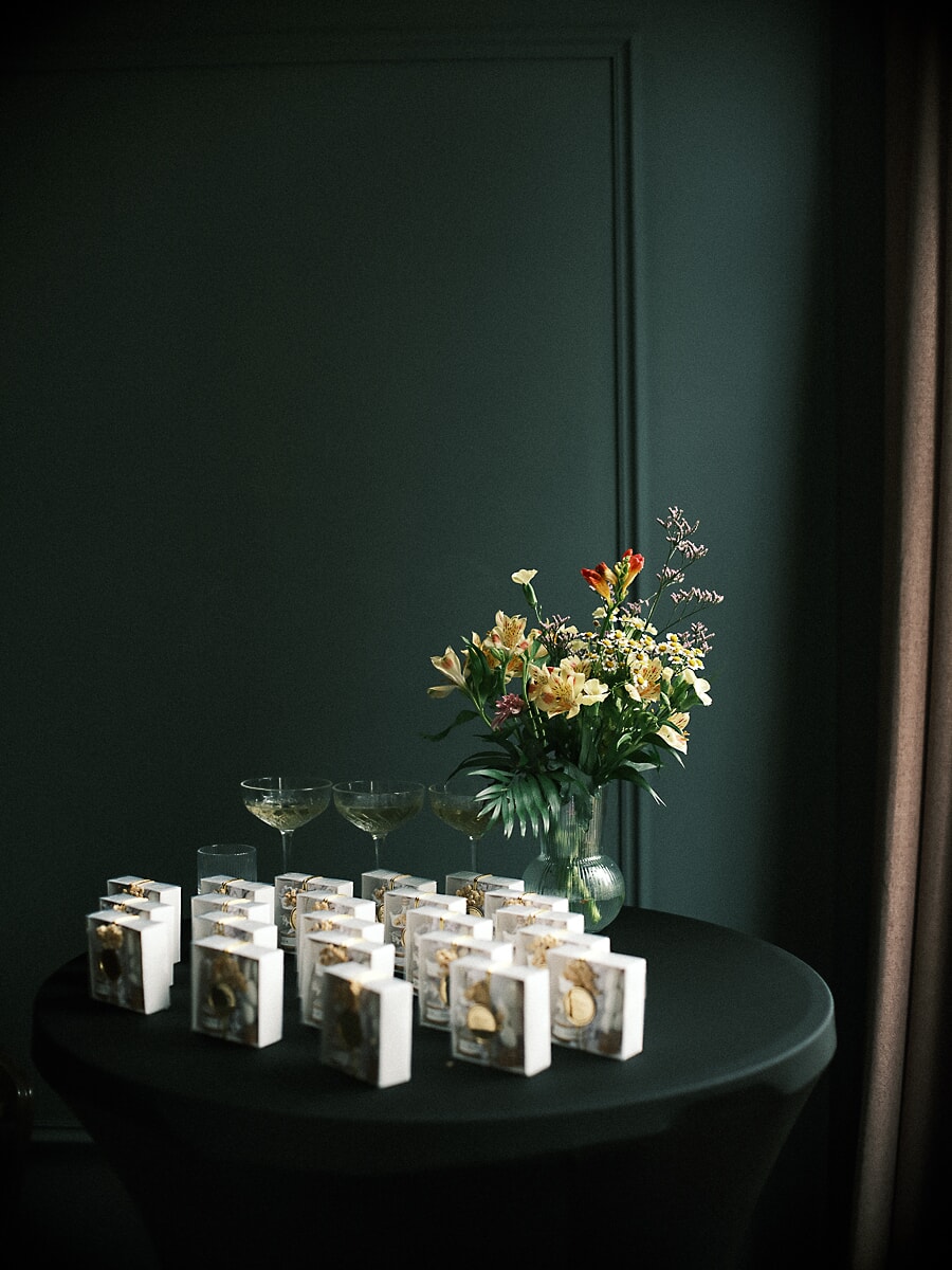 Rijen kleine witte cadeaudoosjes met gouden bedeltjes op een donkere tafel naast coupeglazen en bloemen, productfotografie in Amsterdam